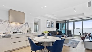 A modern kitchen with a marble backsplash, a dining area with blue chairs, and a large window overlooking the sea. - Luxury Beachfront 1-bed Apartment With Pool & Gym (Gold Coast)