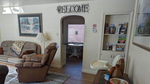 A living room with a brown leather sofa, a framed painting, a 'WELCOME' sign, a no smoking sign, a small table with a lamp, a bookshelf with magazines, and a window with blinds.