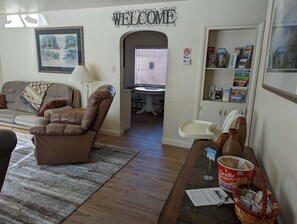 Un salon avec un canapé en cuir marron, un tableau encadré, une pancarte « BIENVENUE », un panneau « INTERDIT DE FUMER », une petite table avec une lampe, une bibliothèque remplie de magazines et une fenêtre avec des stores.