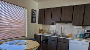 A compact kitchen with dark wooden cabinets, a white countertop, a sink, and a dishwasher. There is a window with blinds, a small dining table with chairs, and a rug on the floor.