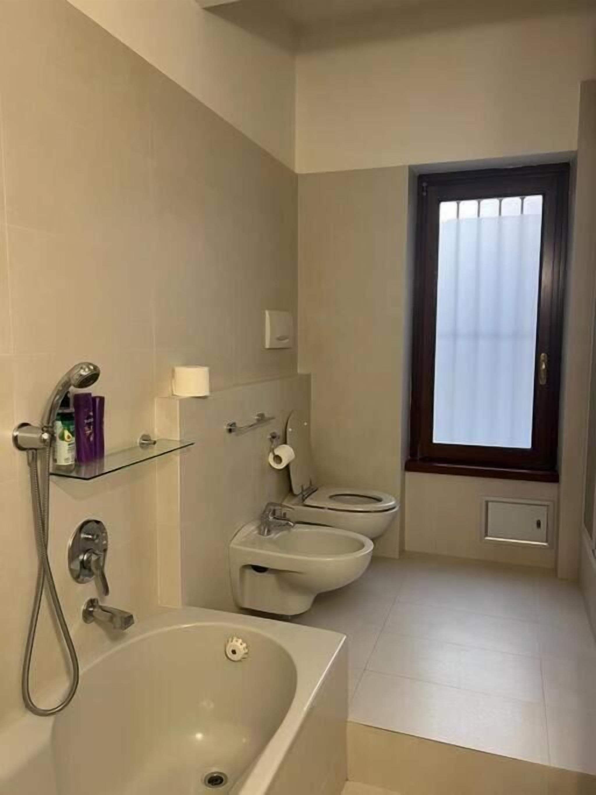 A bathroom with a bathtub, two toilets, a shower, and a window with blinds.