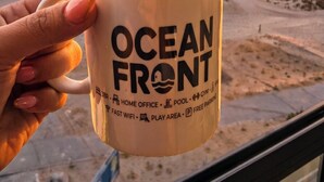A hand holding a mug with 'OCEAN FRONT' text, overlooking a beach with waves and a sunset. - "Oceanfront 3BR, Home Office, Pool, Gym, Sauna, BBQ, Play area and Free Parking" (San Miguel)