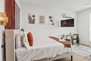 A bedroom with a bed, a desk, a chair, a TV, and a wall-mounted air conditioner. - Designer Suite | Bishop Arts | Gym, Yoga & Games (Dallas)