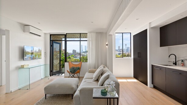 Ein modernes Wohnzimmer mit einer Couch, einem Hocker und einem kleinen Tisch mit einer Vase. An der Wand hängt ein Fernseher, ein Fenster bietet Blick auf die Stadt, und es gibt eine Küchenzeile mit dunklen Schränken und einem Spülbecken.