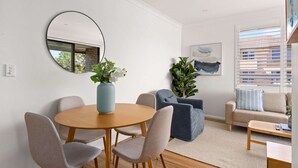 Una sala moderna con una mesa de comedor redonda de madera, sillas tapizadas en gris, un jarrón azul con flores blancas, un sofá azul y un sofá beige con cojines a rayas.