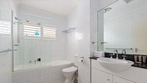 Un baño moderno con ducha envuelta en vidrio, inodoro blanco, lavabo con espejo y ventana con persianas.