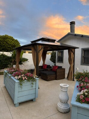 Un espacio de estar al aire libre cubierto, con mobiliario acolchado y jardineras con flores.