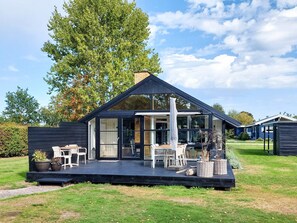 Et moderne hus med en terrasse, udendørs møbler og et stort træ i baggrunden.
