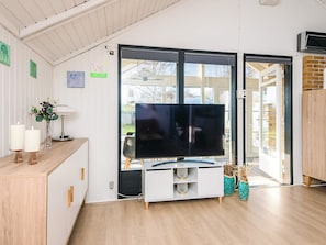 Una sala de estar moderna con un televisor de pantalla plana sobre un mueble blanco, un armario de madera y un ventanal que da al exterior.