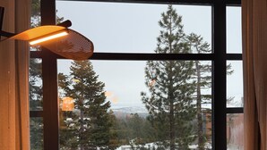 Ein Ecksofa mit Blick auf schneebedeckte Bäume und einen fernen Berg durch ein großes Fenster mit einer gewobenen Leuchte.