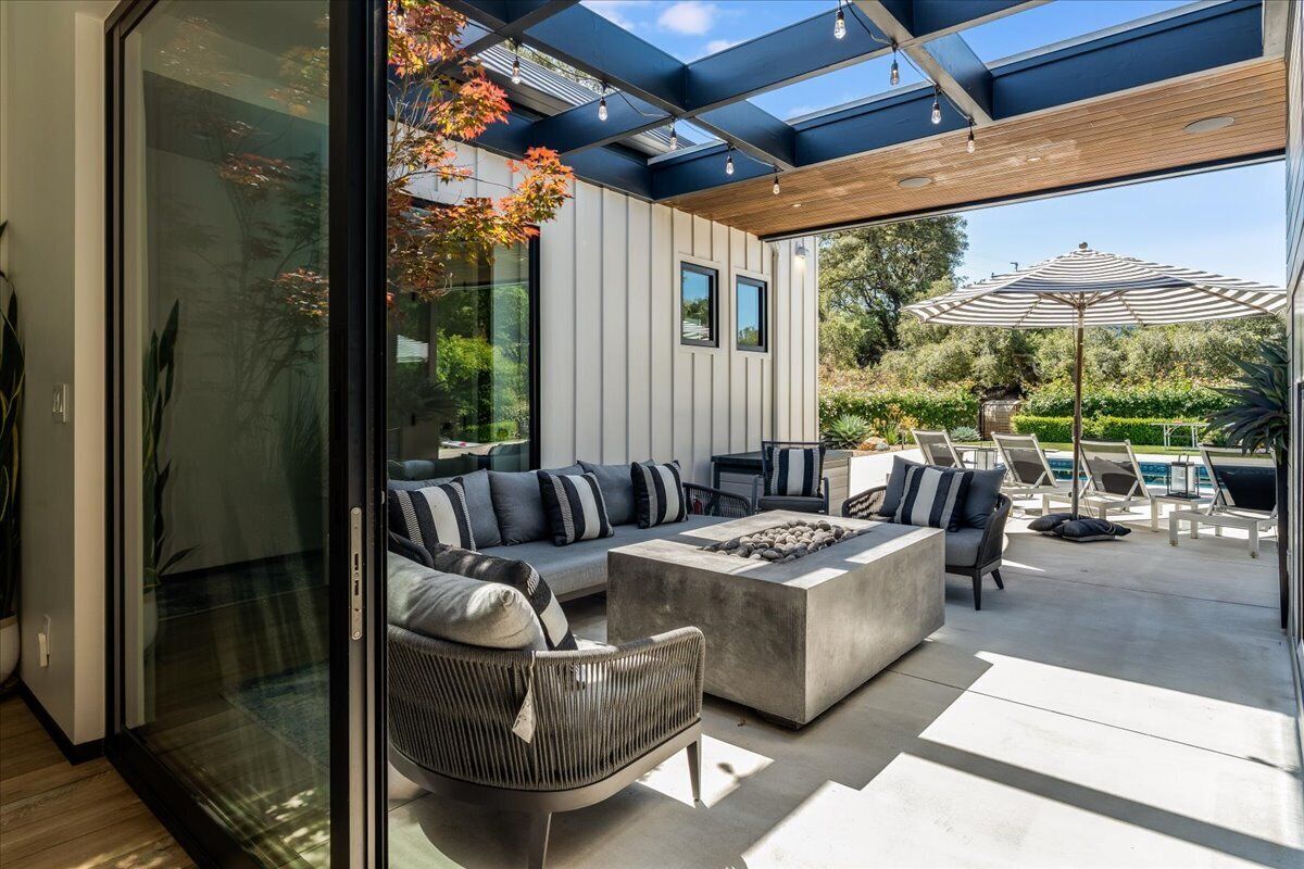 A modern outdoor living space with a glass wall, a built-in fire pit, and a patio area with seating and umbrellas.