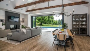 A modern living room with a gray sofa, wooden dining table, and a large window overlooking a pool.