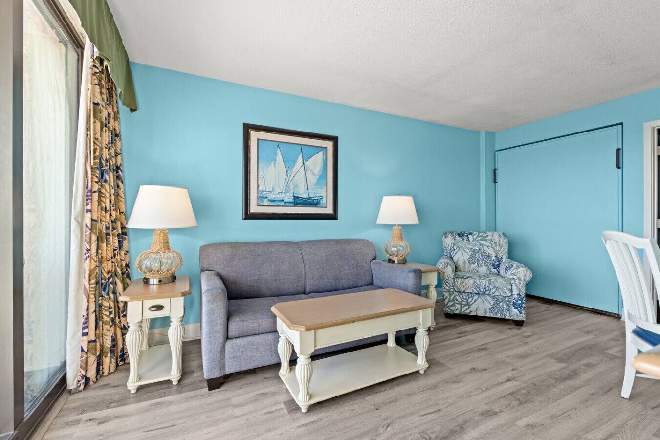 Une pièce avec un mur bleu, un tableau encadré représentant des voiliers, un canapé gris, un fauteuil à motifs, une table basse en bois et deux lampes sur des tables d’appoint.