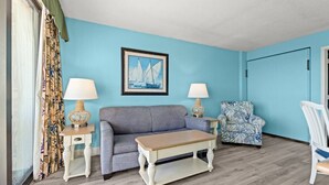 A room with a blue wall, a framed picture of sailboats, a grey sofa, a patterned armchair, a wooden coffee table, and two lamps on side tables. - Beachfront End Unit Condo with Pools and Hot tubs (Myrtle Beach)