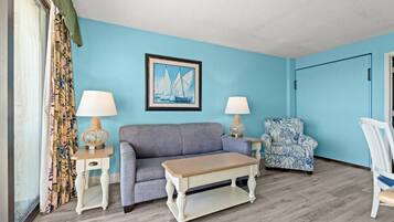 Une pièce avec un mur bleu, un tableau encadré représentant des voiliers, un canapé gris, un fauteuil à motifs, une table basse en bois et deux lampes sur des tables d’appoint.