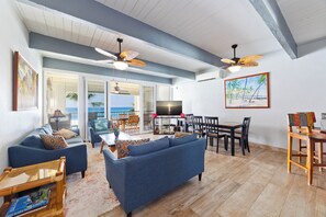 Ein großzügiges Wohnzimmer mit einem blauen Sofa, einem Holzesstisch und Stühlen. Es gibt Deckenventilatoren, einen Fernseher und ein Bild mit einer Strandansicht.