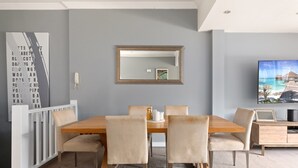 Un coin repas avec une table en bois et des chaises de couleur beige, un téléphone blanc, un miroir encadré et un téléviseur fixé au mur.