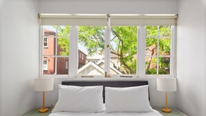 Un lit bien fait, avec du linge de lit blanc, deux lampes de chevet et une vue sur un bâtiment en briques par une grande fenêtre.