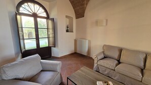 Un soggiorno con un divano, una poltrona, un tavolino in legno, un termosifone e una finestra con porta aperta.