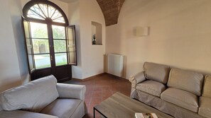 A living room with a sofa, a chair, a wooden coffee table, a radiator, and a window with an open door. - Historic House Casa Magnolia - Castello di Vecchienna, 30mn from the sea (Toscana)