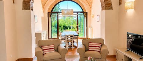 A living room with two armchairs, a coffee table with books and a vase, a TV stand, and a large arched brick ceiling.