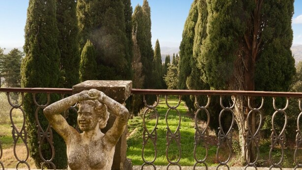 Una statua in pietra raffigura una donna che si tiene la testa, con sullo sfondo alti cipressi e un cielo sereno.