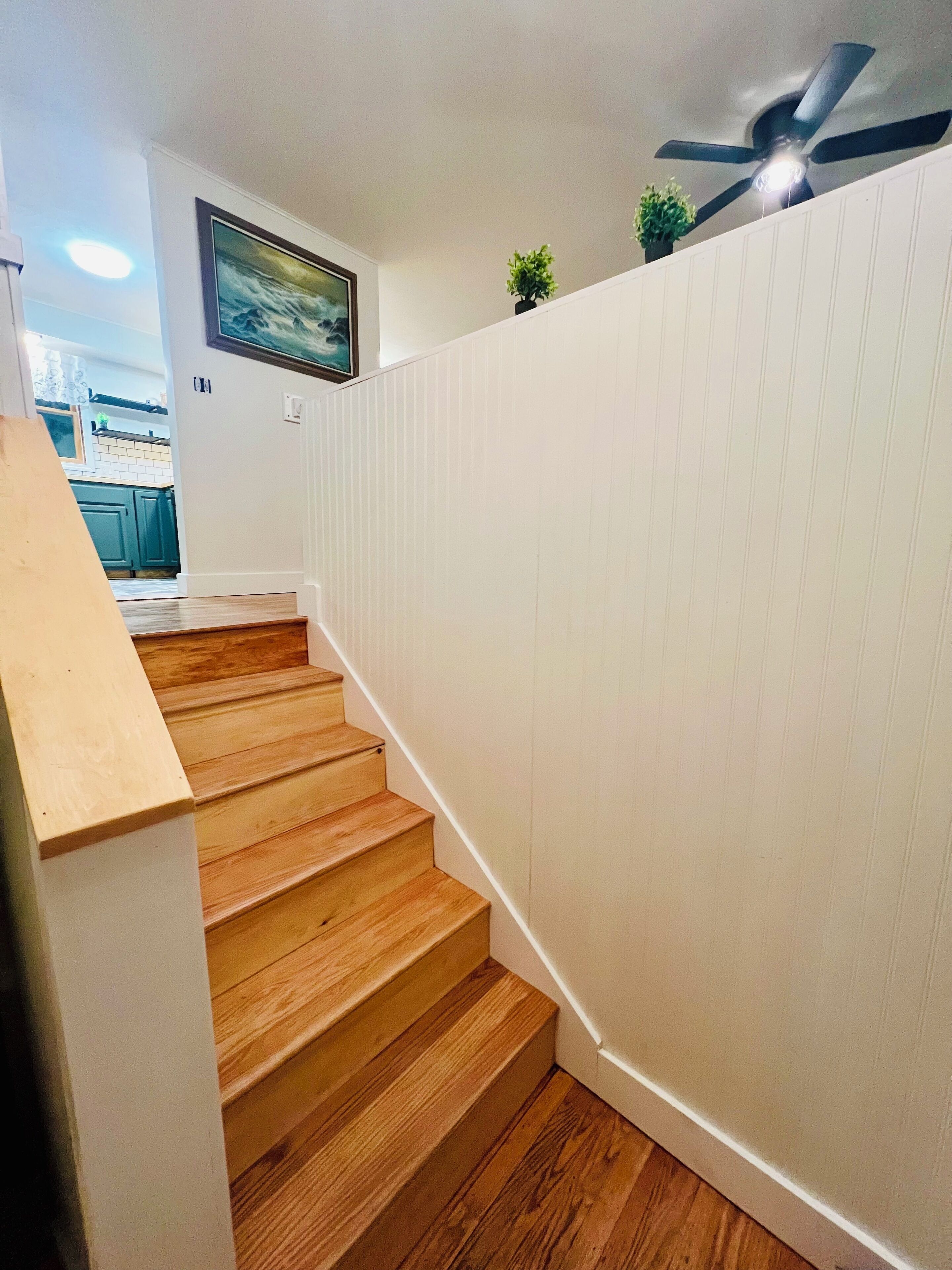 Un escalier en bois, avec des lambris blancs sur les murs, mène à une cuisine dotée de meubles verts et d’un ventilateur de plafond.