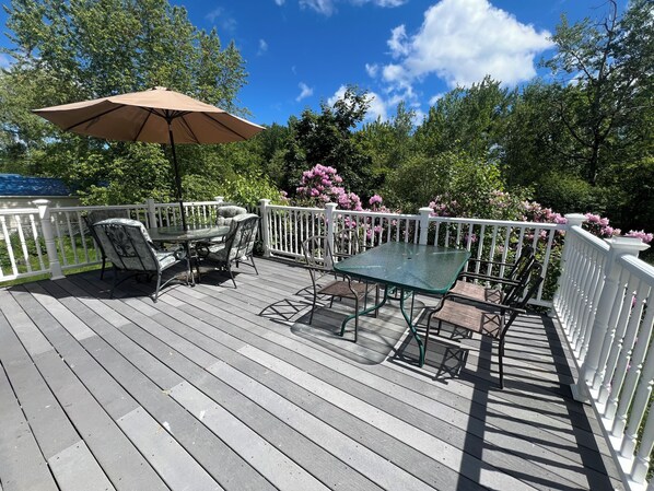 A wooden deck with a glass table and chairs, surrounded by a white railing and lush greenery.