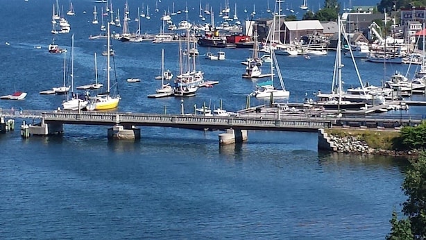 Uma marina com vários barcos atracados, uma ponte sobre a água e edifícios ao fundo.