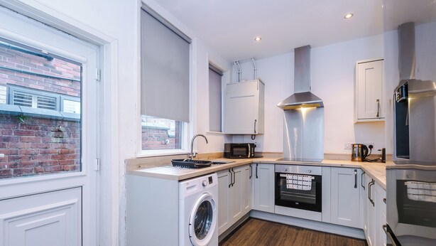 A compact kitchen with a washing machine, microwave, oven, and a large window with a view of a brick building. - Walkden, Worsley 4 Bedroom House w/ Free Parking (Worsley)