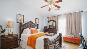 Un dormitorio con una cama de madera, una mesita de noche, un escritorio, una silla, un ventilador de techo, un espejo y una ventana con cortinas.