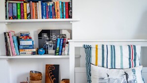 Une chambre avec un lit blanc, des oreillers à rayures et une bibliothèque remplie de livres et d’objets décoratifs.