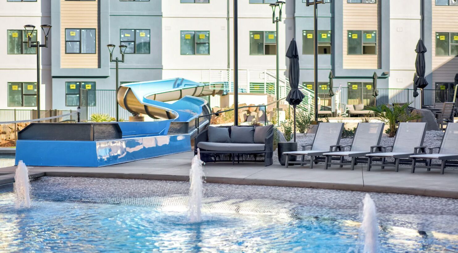 Zona de piscina con sillones reclinables, un tobogán de agua azul y un edificio con múltiples ventanales al fondo.