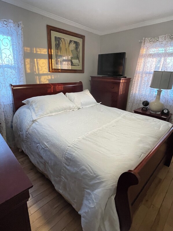 Une chambre à coucher avec un lit en bois, un téléviseur posé sur une commode en bois, un tableau encadré accroché au mur et une fenêtre avec des rideaux.