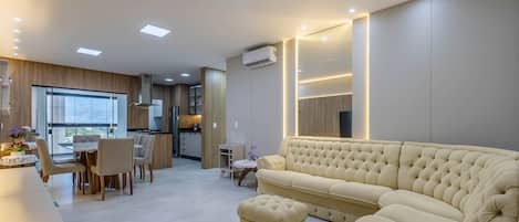 Un salon moderne avec un canapé beige, un coin repas comprenant une table et des chaises, et une cuisine dotée de meubles en bois et d’un four encastré.