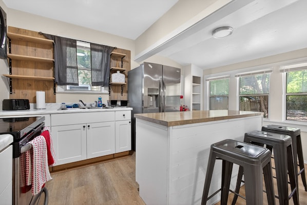 A modern kitchen with white cabinets, a stainless steel refrigerator, wooden shelves, and a large island with stools.
