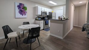A compact kitchen with white cabinets, a microwave, and a refrigerator. There is a dining area with a round table and chairs, and a kitchen island with a sink.