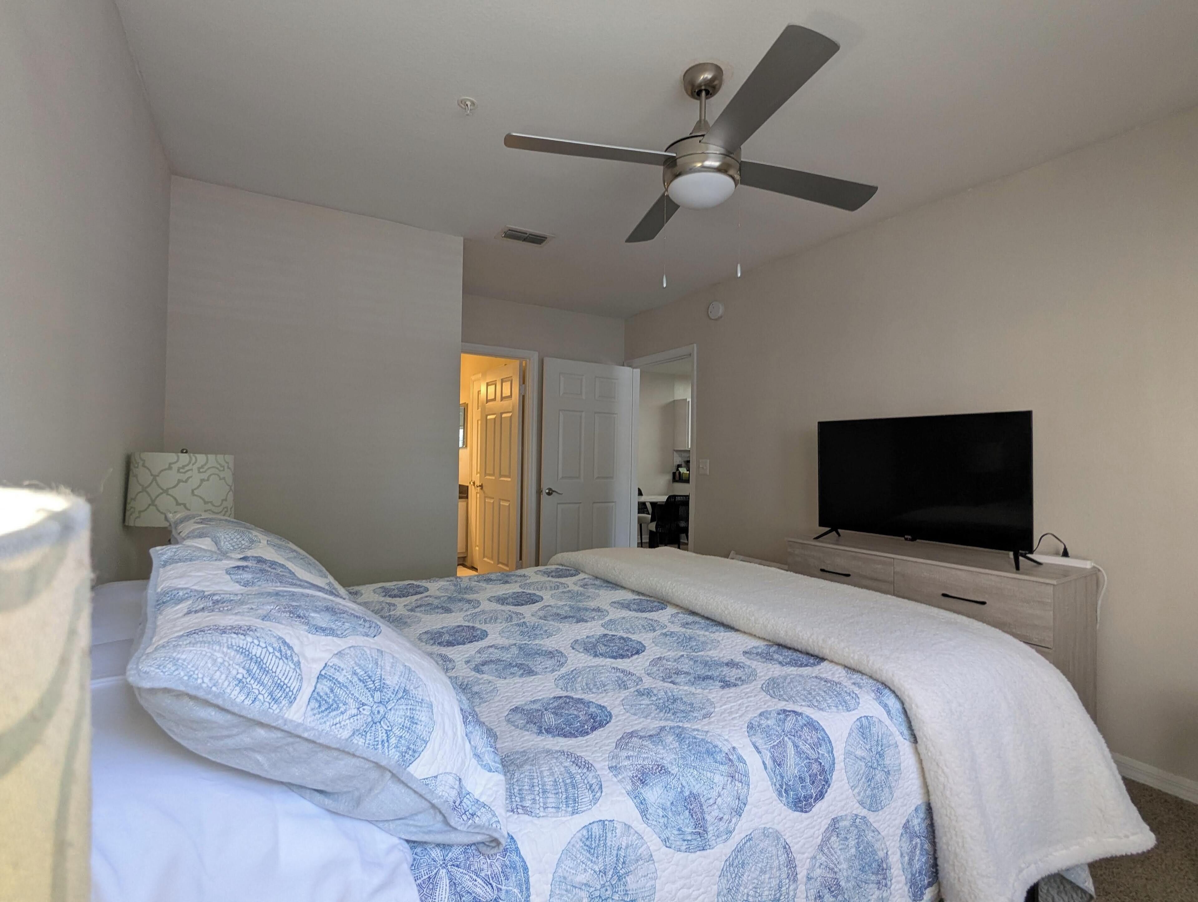 A bedroom with a bed, a ceiling fan, a TV, and a door leading to a bathroom.
