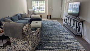 A living room with a sectional sofa, a coffee table, a TV on a stand, and a large window with blinds.