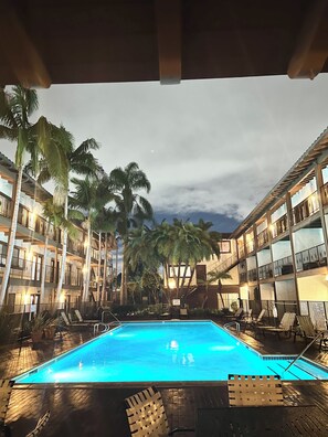 Área de piscina com espreguiçadeiras e palmeiras, cercada por edifícios com vários andares e sacadas.