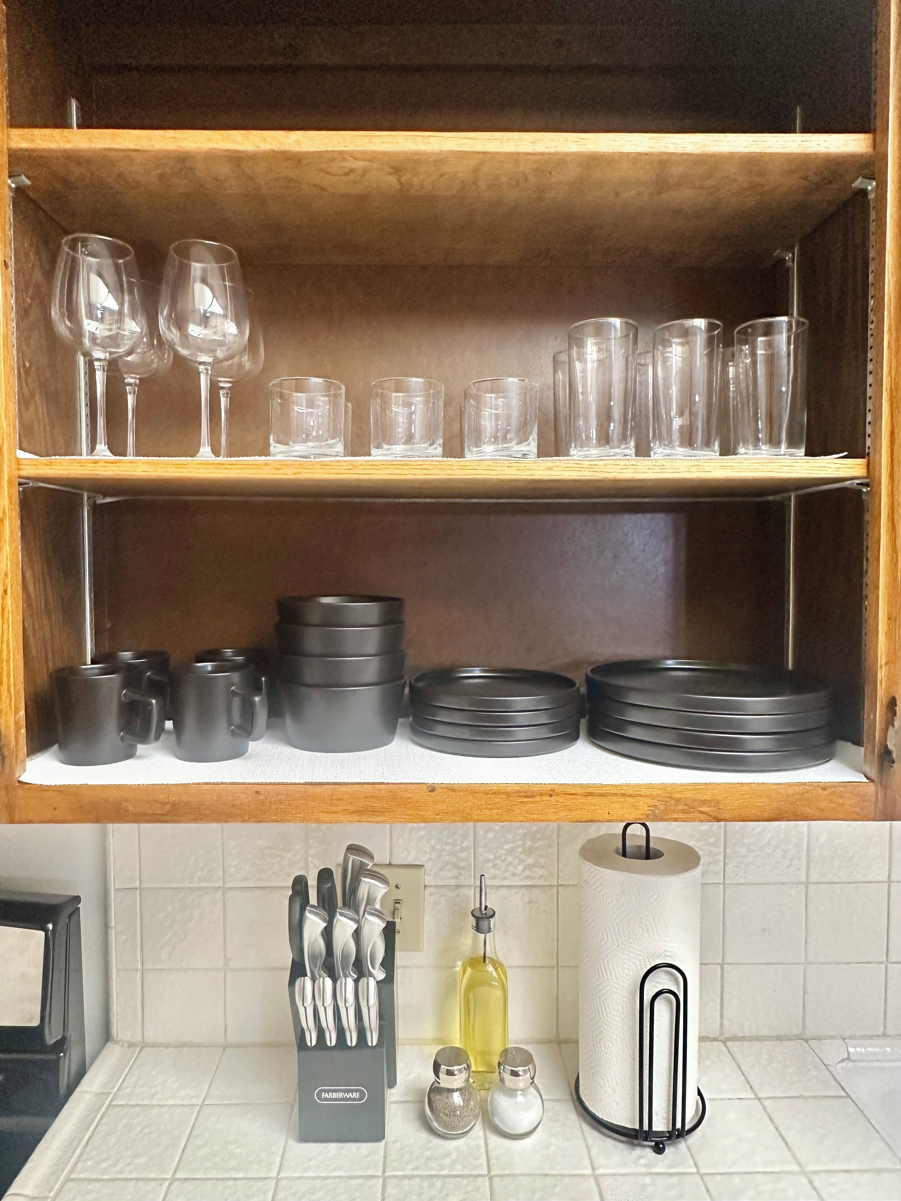 Una estantería de cocina con copas de vino, tazas y platos. Abajo, hay utensilios, una botella de aceite, saleros y pimenteros, y un dispensador de toallas de papel.