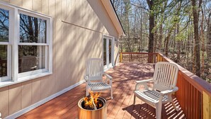 Una terraza de madera con dos sillas Adirondack blancas, un brasero y vistas del bosque circundante.