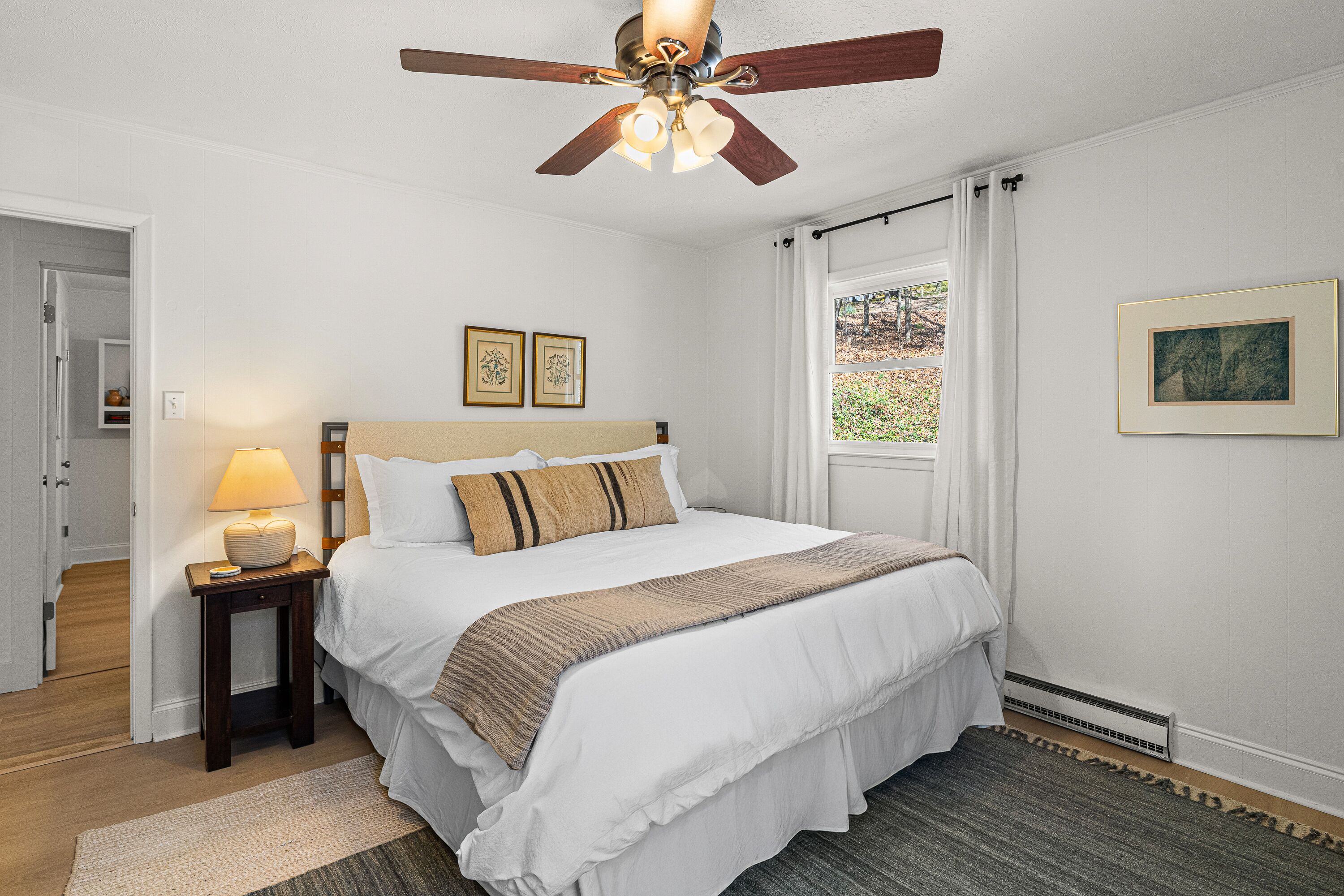 A bedroom with a bed, a ceiling fan, a nightstand, a lamp, a window with curtains, and framed pictures on the wall.