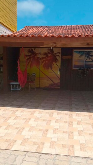 A building with a tiled roof, a mural of palm trees and a sunset, a small table with a chair, and a wooden door. - Jô's Corner (Florianópolis)