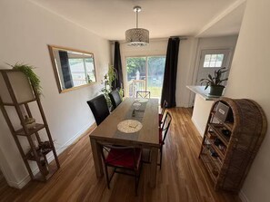 A dining area with a wooden table, chairs, and a hanging light fixture. There is a window with curtains, a plant on a shelf, and a wicker chair. - Giulietta & Romeo’s home. Backyard, Free parking (1st floor) (Niagara Falls)