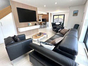 A modern living room with a black leather sofa, a coffee table, and a flat-screen TV mounted on a wooden wall. There is a dining area with a table and chairs in the background. - VILLA DETENTE A BISCA (BISCARROSSE)