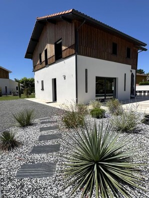 A modern building with a white facade and wooden upper structure, surrounded by gravel and plants. - VILLA DETENTE A BISCA (BISCARROSSE)