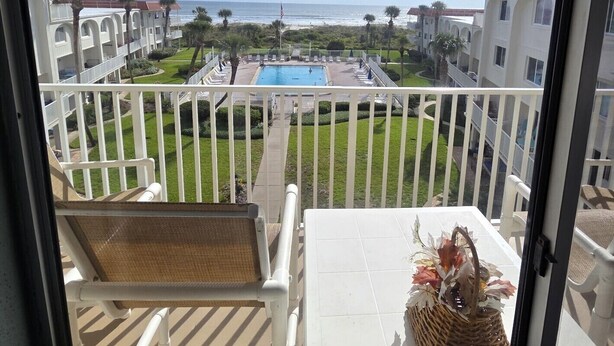 En balkong med utsikt över en pool och havet, omgiven av byggnader med välvda balkonger och palmer.