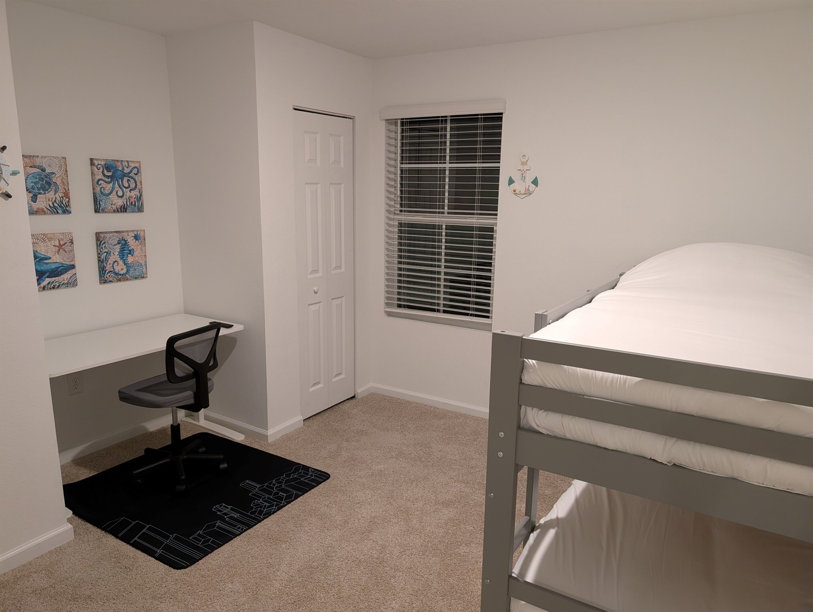 A bedroom with a bunk bed, a desk with a chair, a window with blinds, and four wall-mounted artwork.