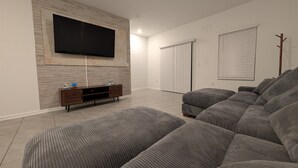 Un salón con un televisor de pantalla plana grande montado en una pared de madera, un sofá seccional gris, un mueble bajo de madera y un ventanal corredizo que da a un balcón.